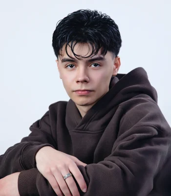 A close-up portrait of a young man with dark, styled hair and a silver ring on his hand, wearing a brown hoodie against a white background.