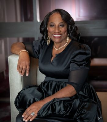 A portrait of an older woman with styled dark hair, smiling and wearing a black satin dress, gold jewelry, and a pearl necklace while sitting on a light-colored armchair.