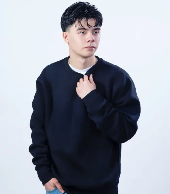 A portrait of a young man with dark, styled hair wearing a black sweatshirt over a white t-shirt, adjusting his collar against a white background.