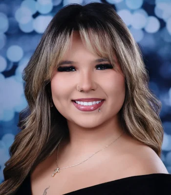 A close-up headshot of a smiling woman with layered blonde and brown hair, a nose ring, lip piercing, and a gold cross necklace against a blue bokeh background.