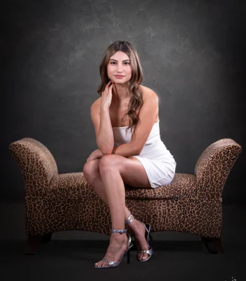 A portrait of a woman with long brown hair wearing a white strapless dress and silver ankle-strap heels, sitting on an animal-print bench against a dark textured background.