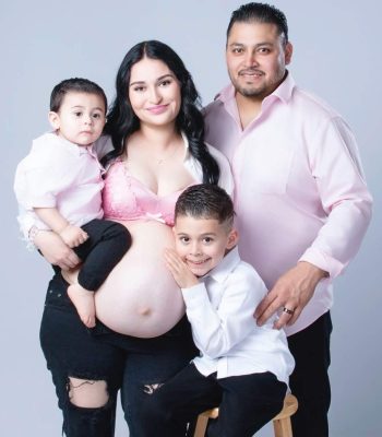 pregnant mother in white lace top, husband, and young son sitting together against a white studio backdrop